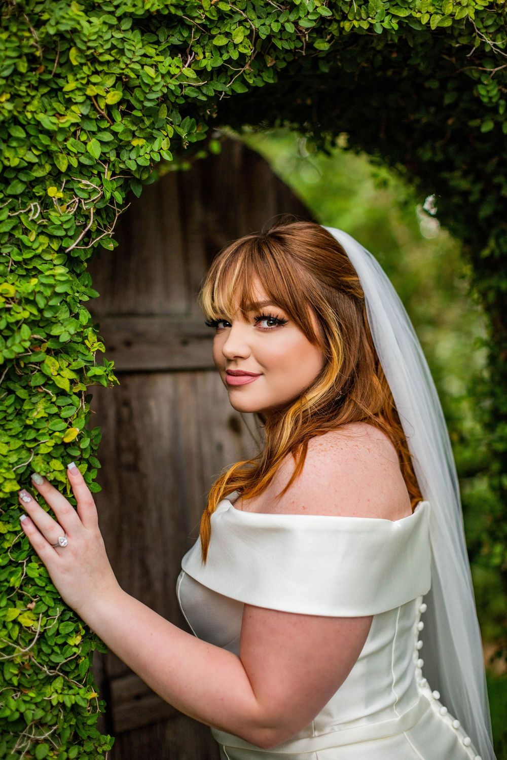 Bride in off-shoulder wedding dress poses gracefully among green garden leaves.