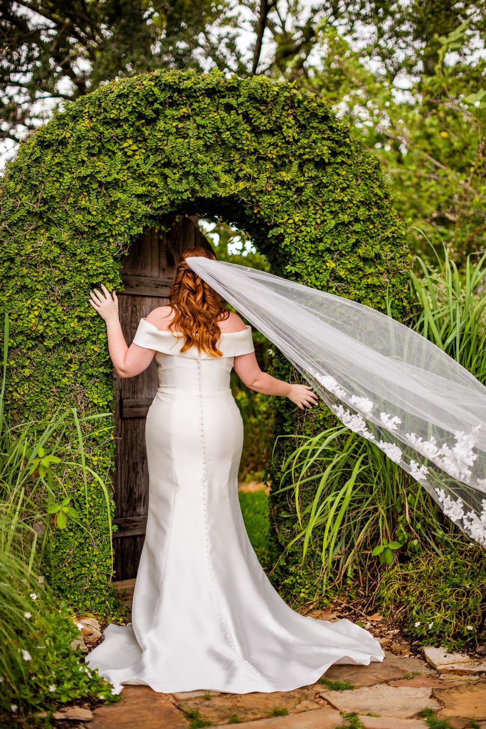Flowing veil catches the breeze as bride poses in garden landscape.