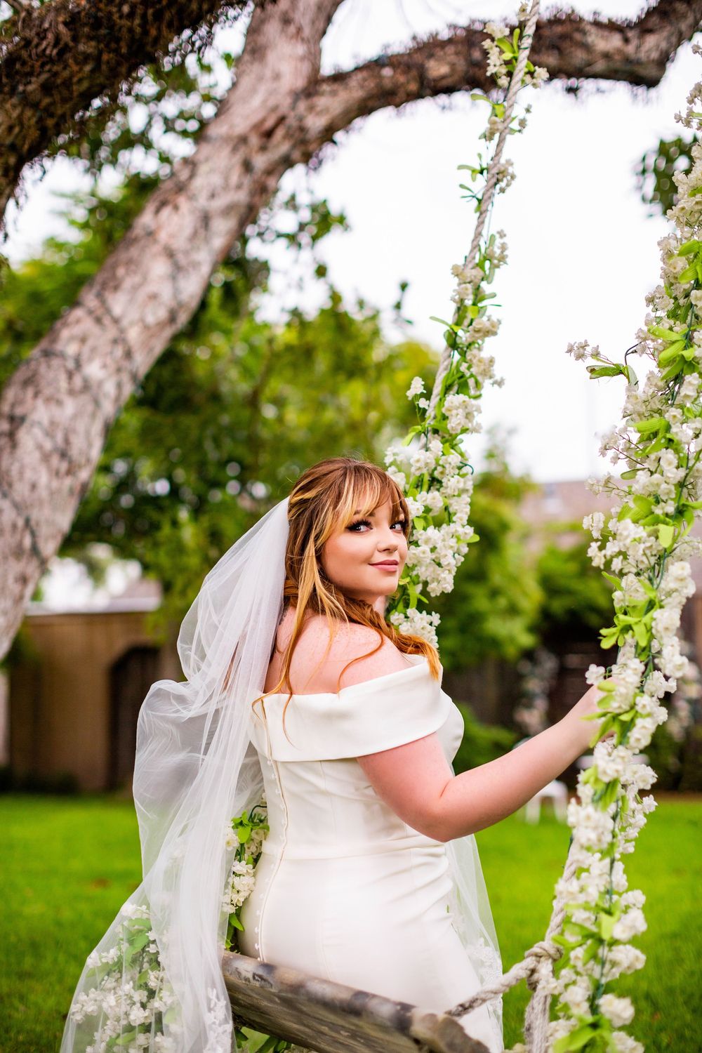 Bride in white dress poses with blooming white flowers in garden setting.