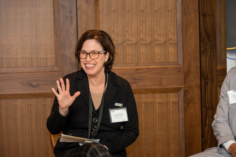A person in glasses and dark clothing stands in front of wooden paneled walls while gesturing with their hand.