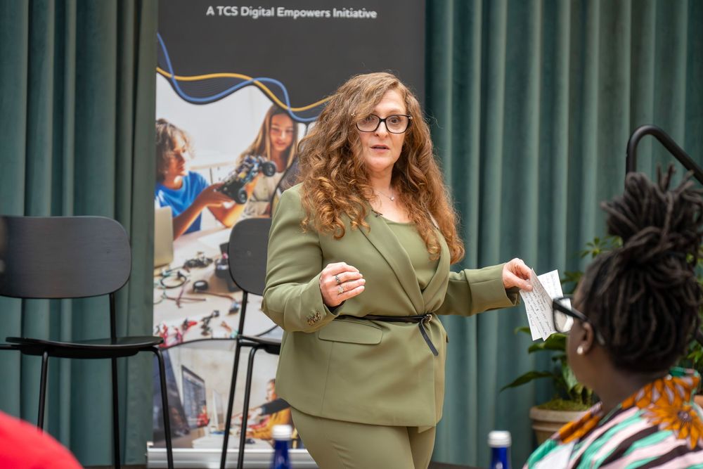 Woman in olive green sweater gestures expressively while speaking at a professional event.