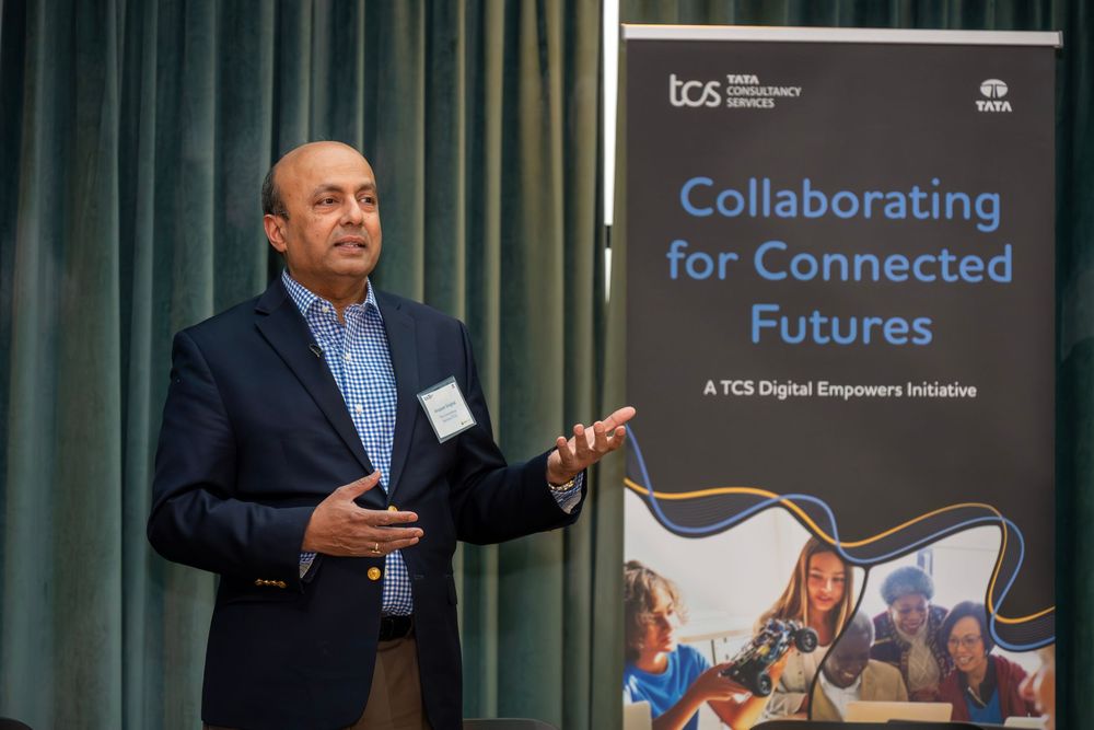 Professional speaker gestures while presenting at a business conference with blue banners and digital displays in background.