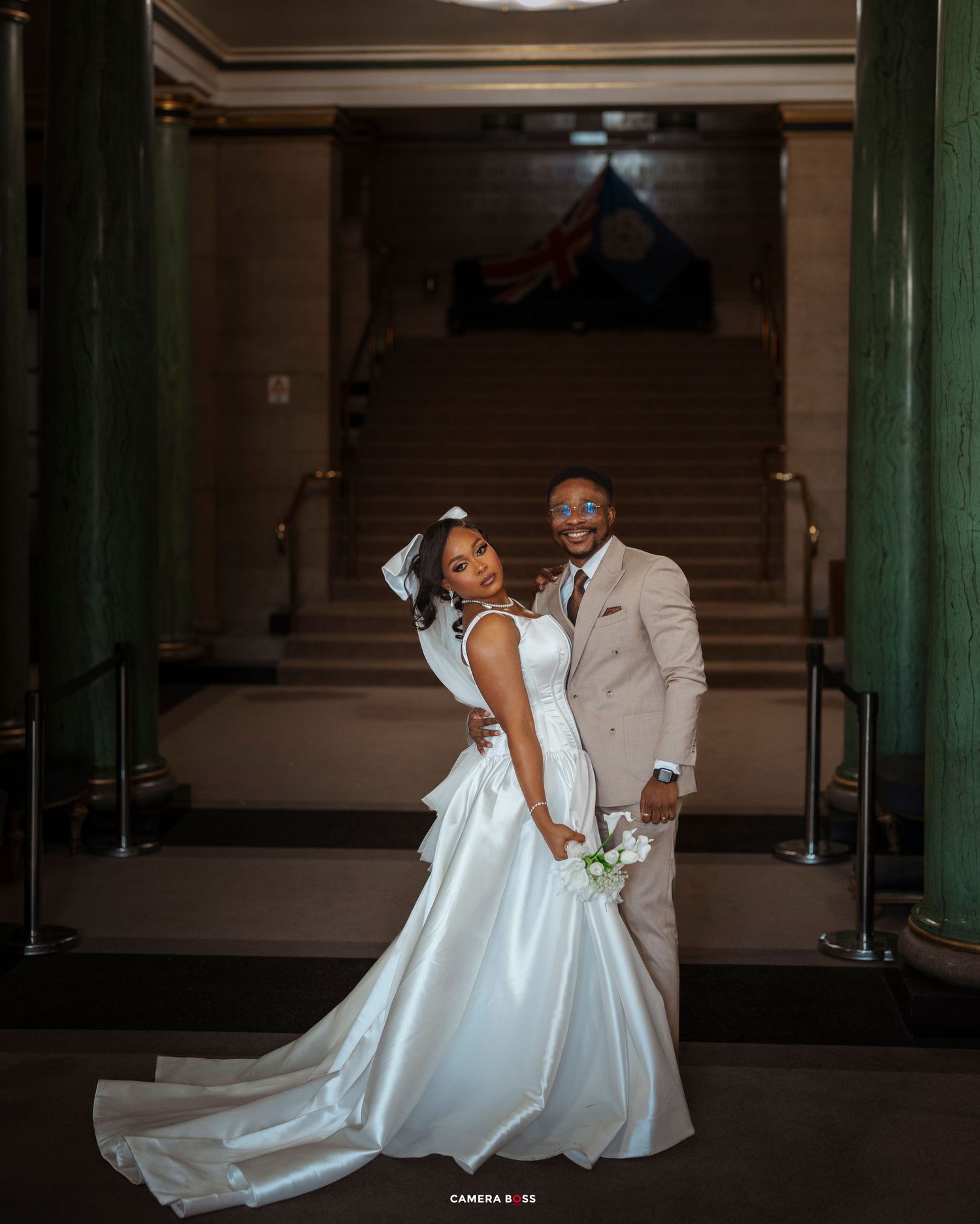 A couple in wedding attire pose elegantly in a grand hallway with green columns and dramatic lighting.