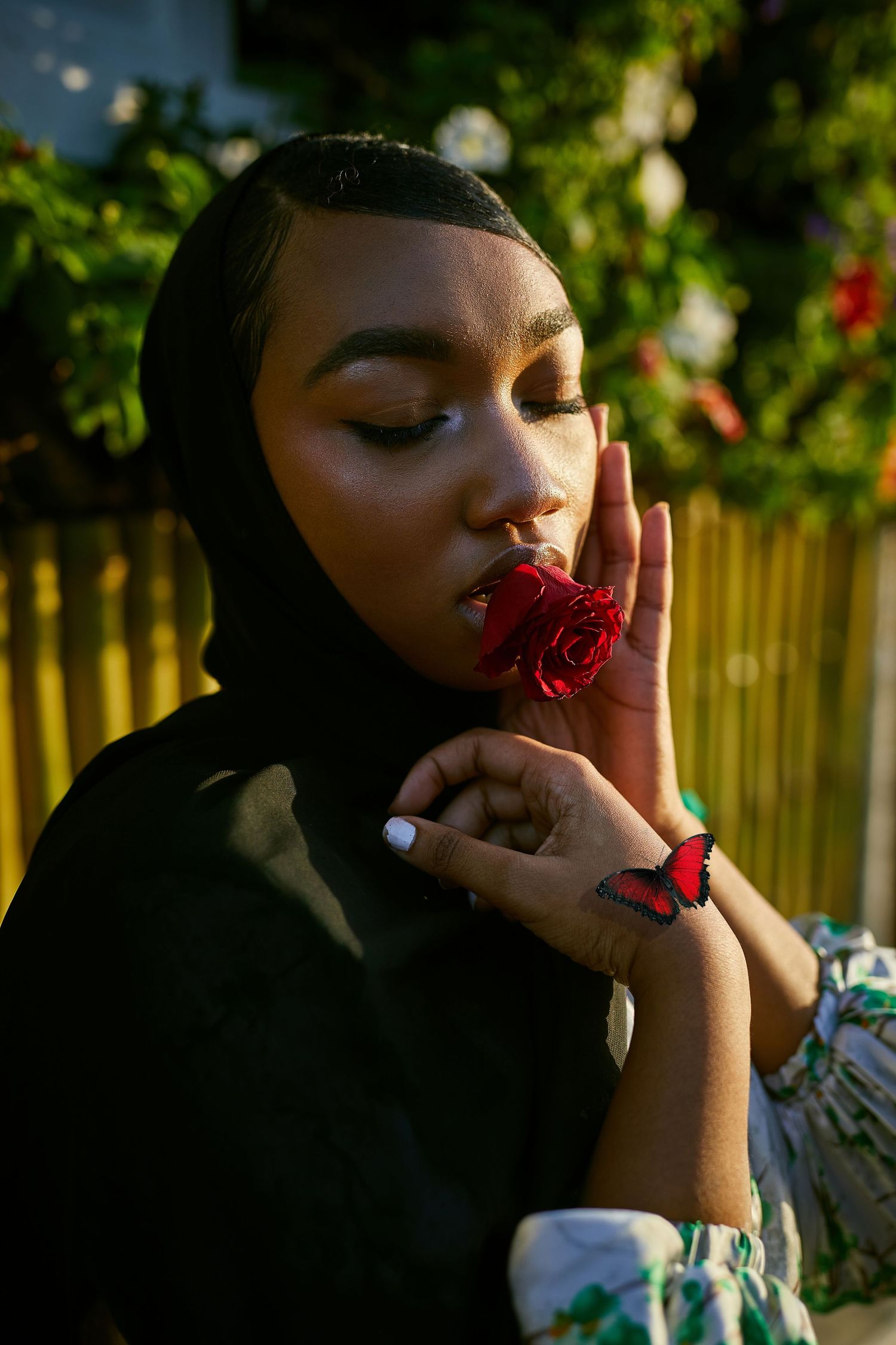 Person in black hijab holding red rose against garden background in golden hour lighting.