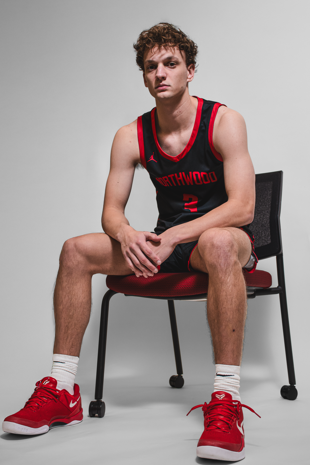 Team Media Day Photo of a basketball athlete wearing a red and black basketball jersey sitting on a chair against white background.
