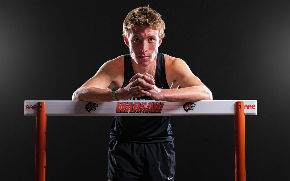 Team Media Day Photo of track athlete leaning over hurdle in dramatic black and white portrait against dark background - Warsaw, Indiana