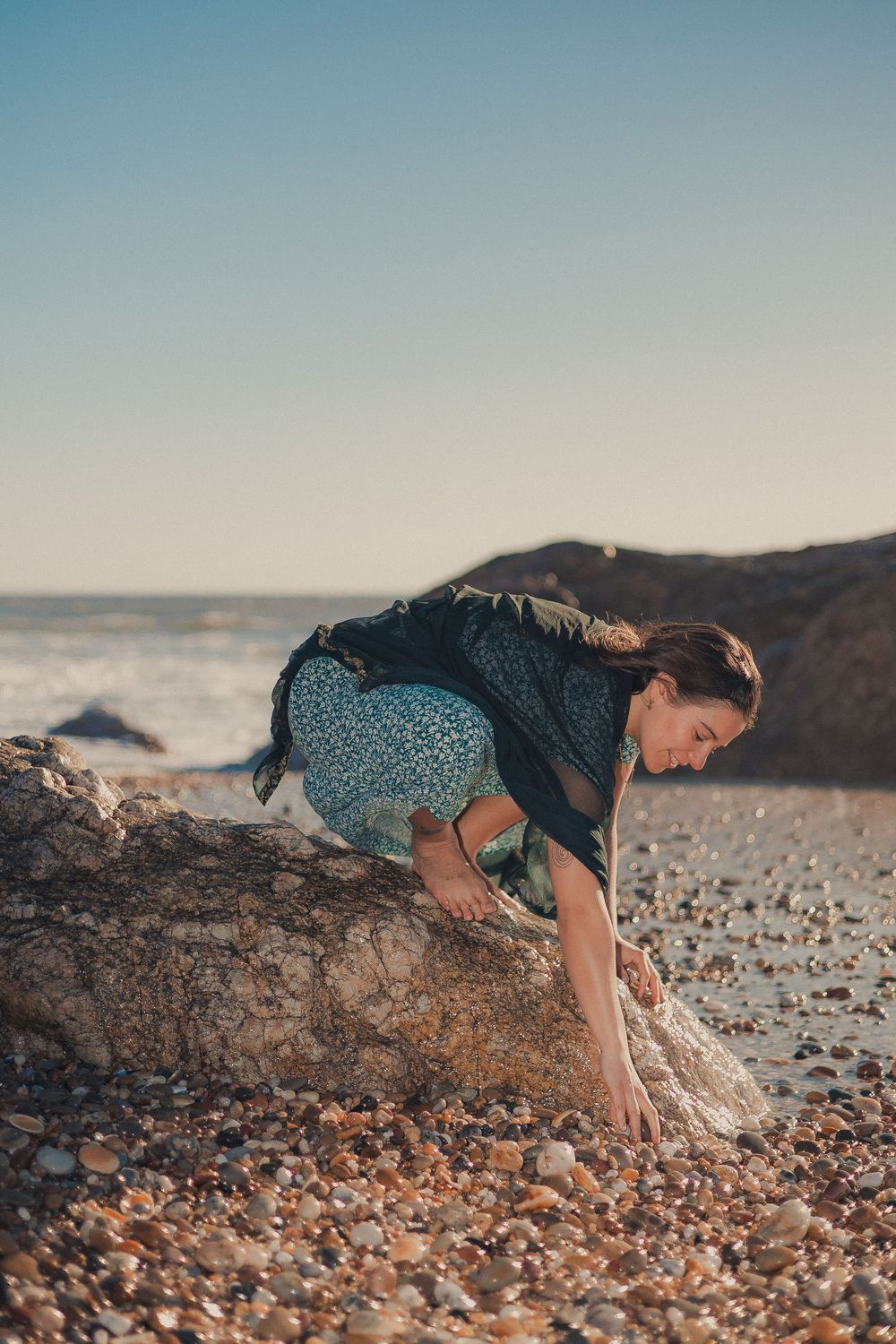 Figure exploring rocky beach landscape in floral dress during golden hour.