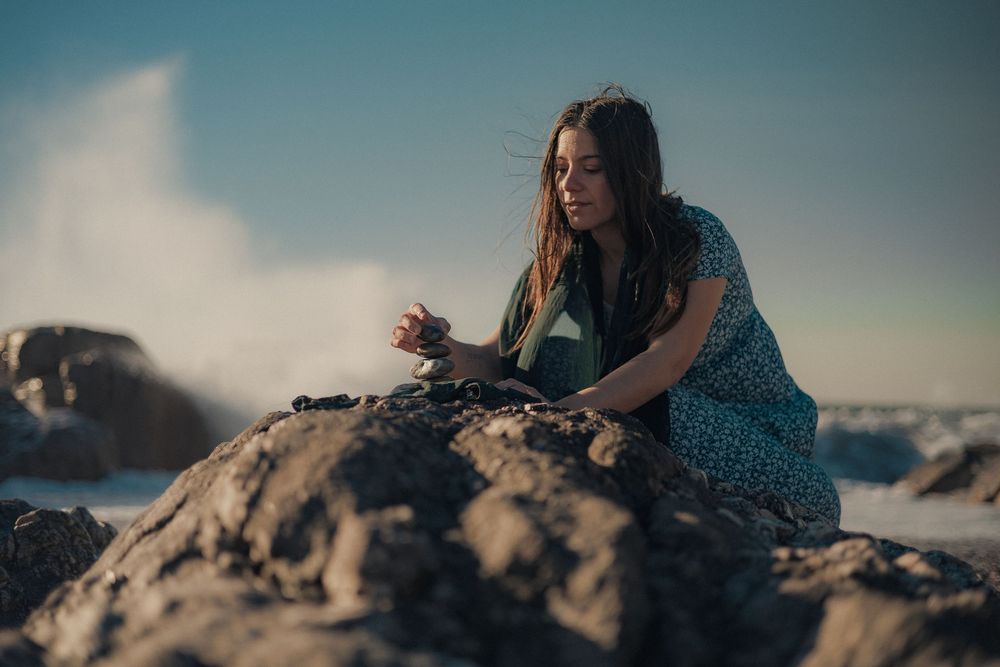 Figure in blue dress sits contemplatively on rocky coastal cliffs at sunset.