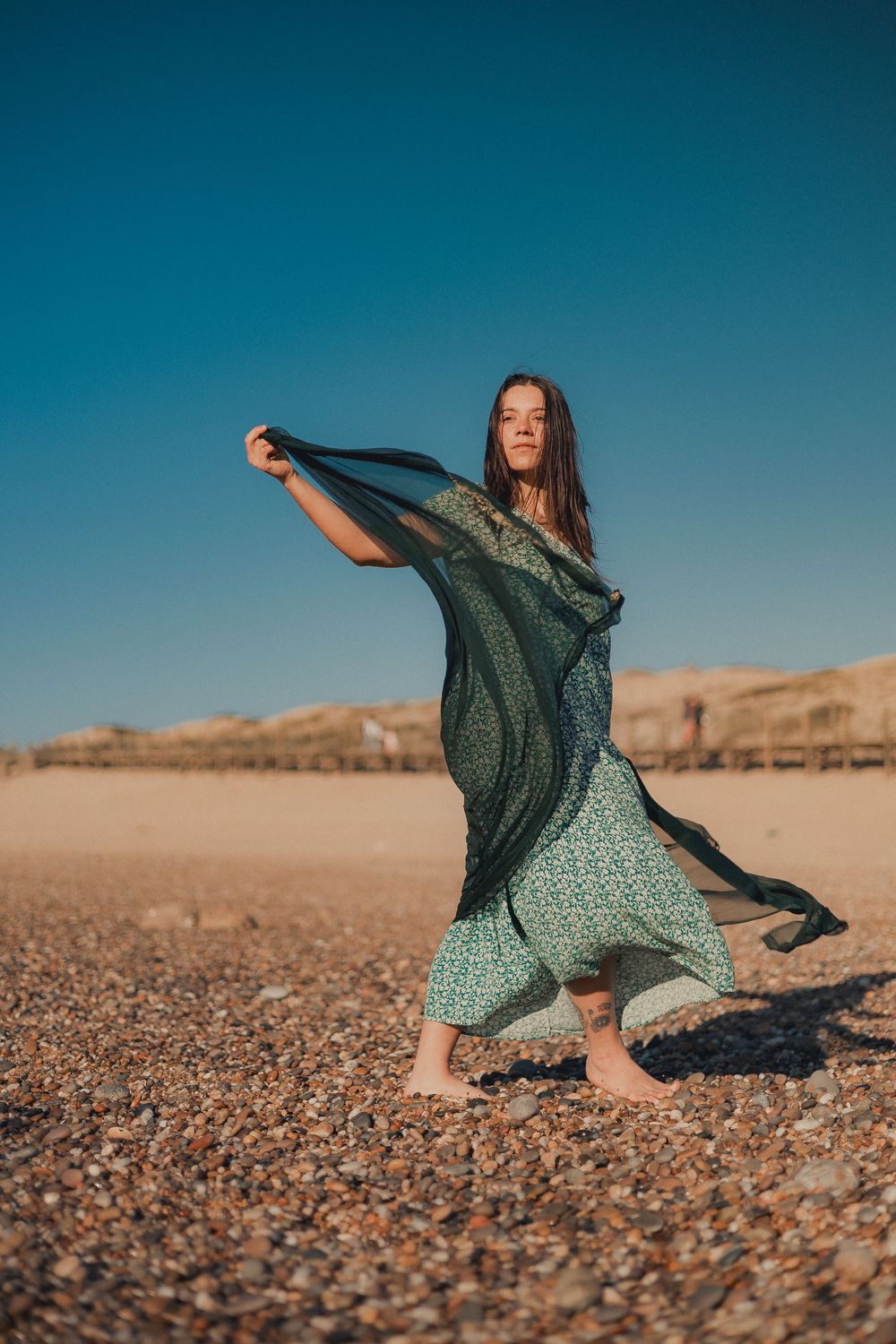 Flowing dress catches wind as figure dances on pebbled beach at sunset.