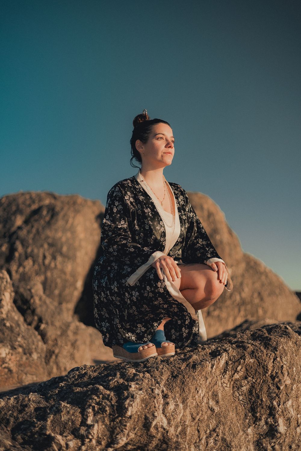 Person in dark floral dress sits thoughtfully on rocky outcrop against blue sky.