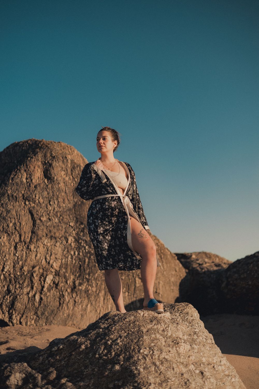 Figure sits atop large rock formations in desert landscape during sunset.