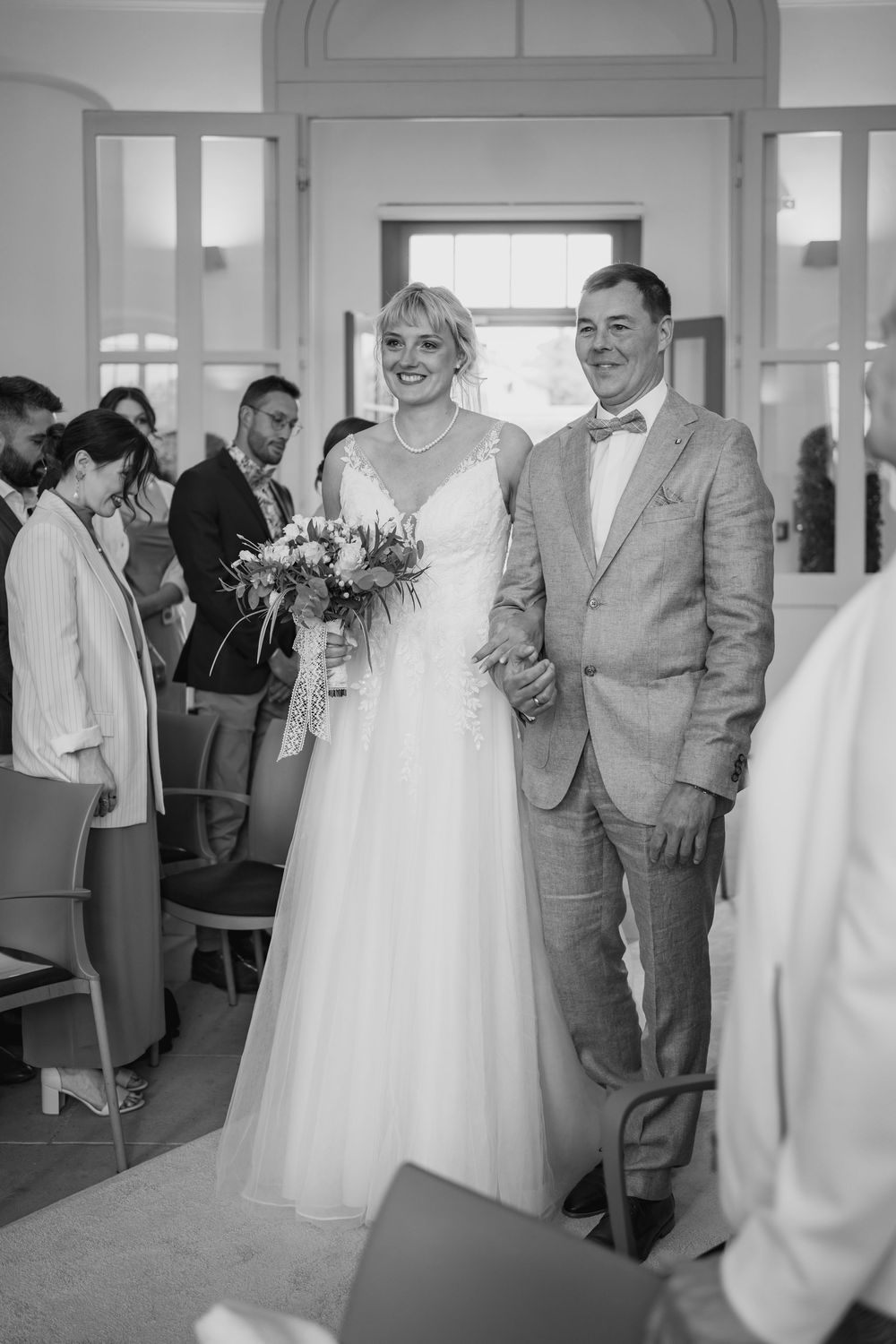 A black and white photograph of a wedding ceremony in a classic interior setting with a couple walking down the aisle.