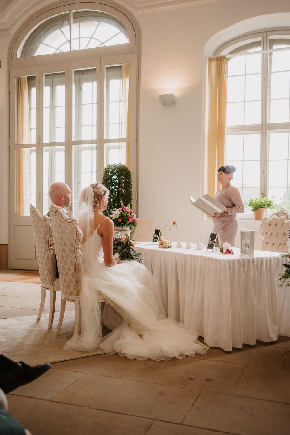 Wedding ceremony in an elegant room with tall arched windows, white decor and a draped table setting.