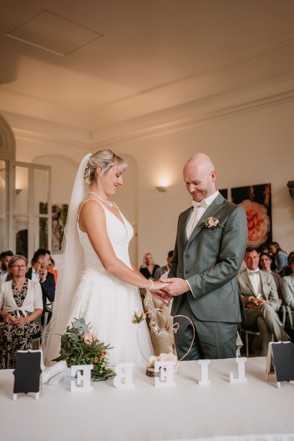 A couple exchanges rings during their wedding ceremony at an elegant indoor venue with soft lighting and floral decor.