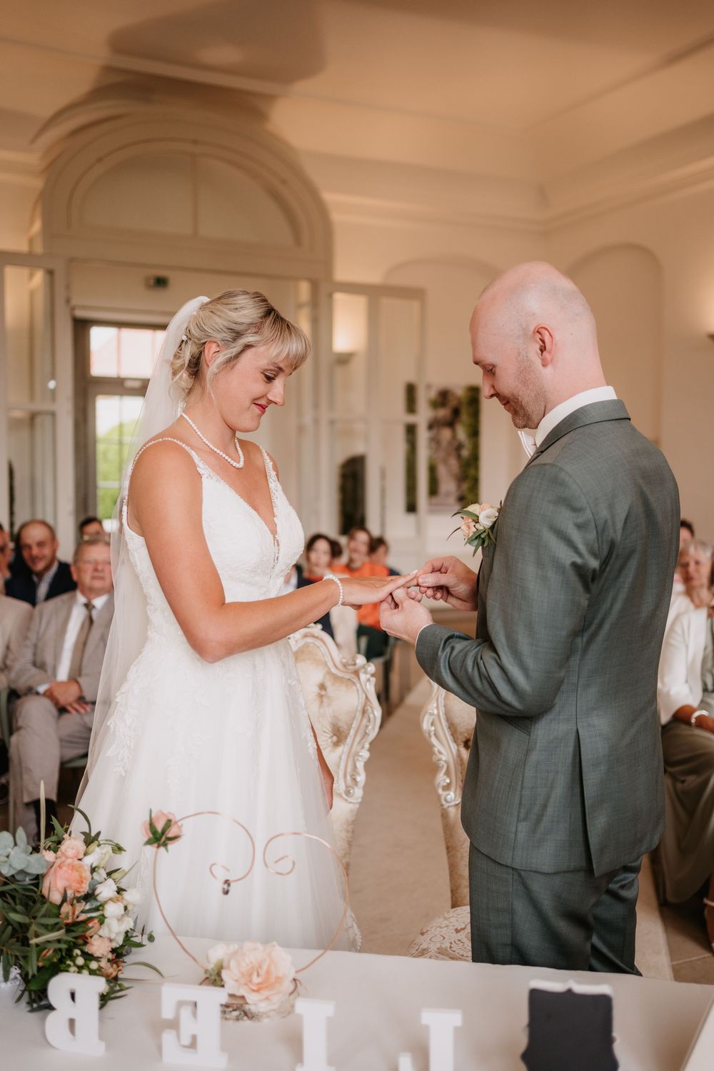 A romantic wedding ceremony in an elegant room with floral decorations and guests watching the exchange of rings.