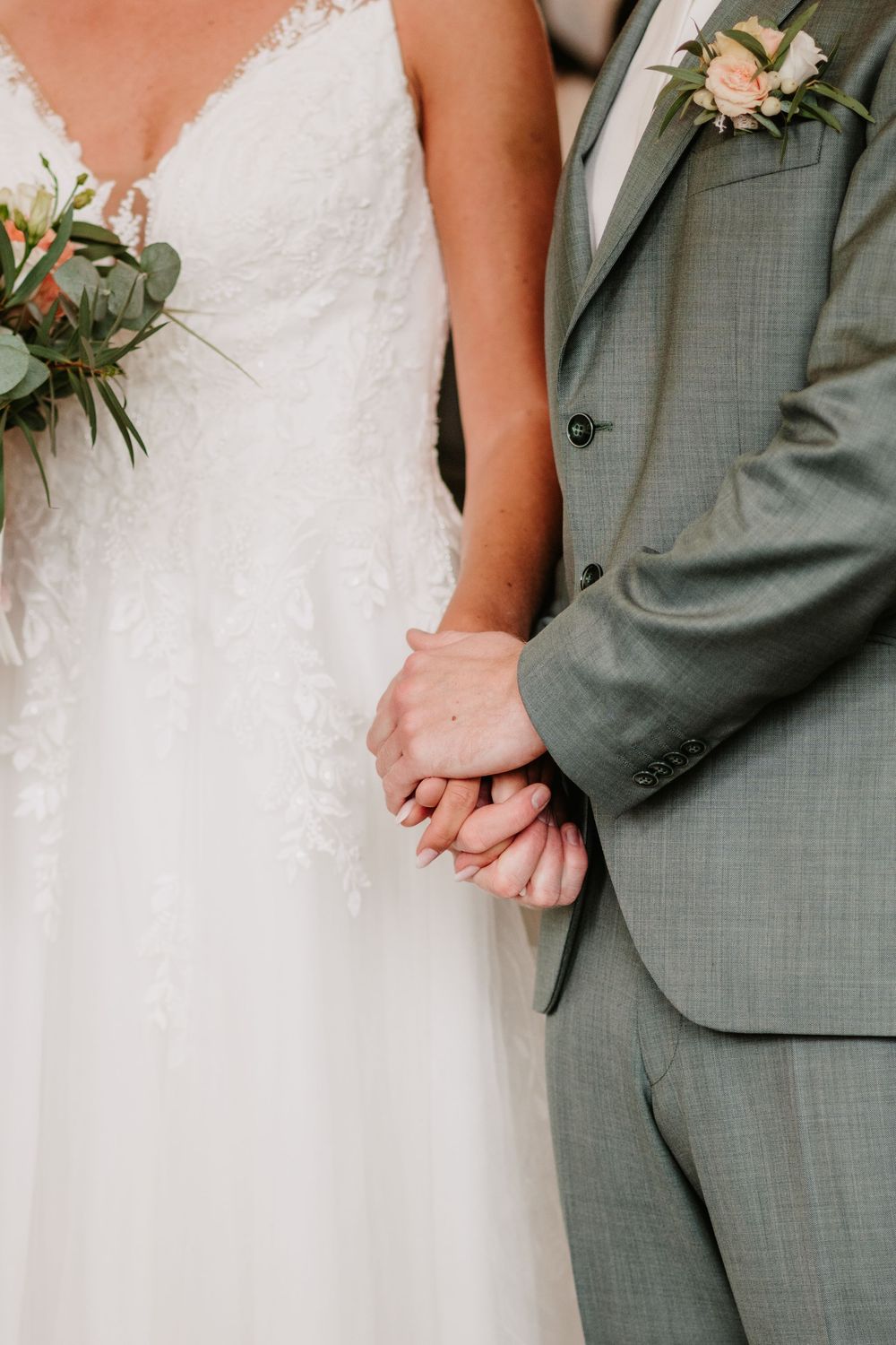 Close-up of newlyweds holding hands, showing white lace wedding dress and gray suit with floral boutonniere.