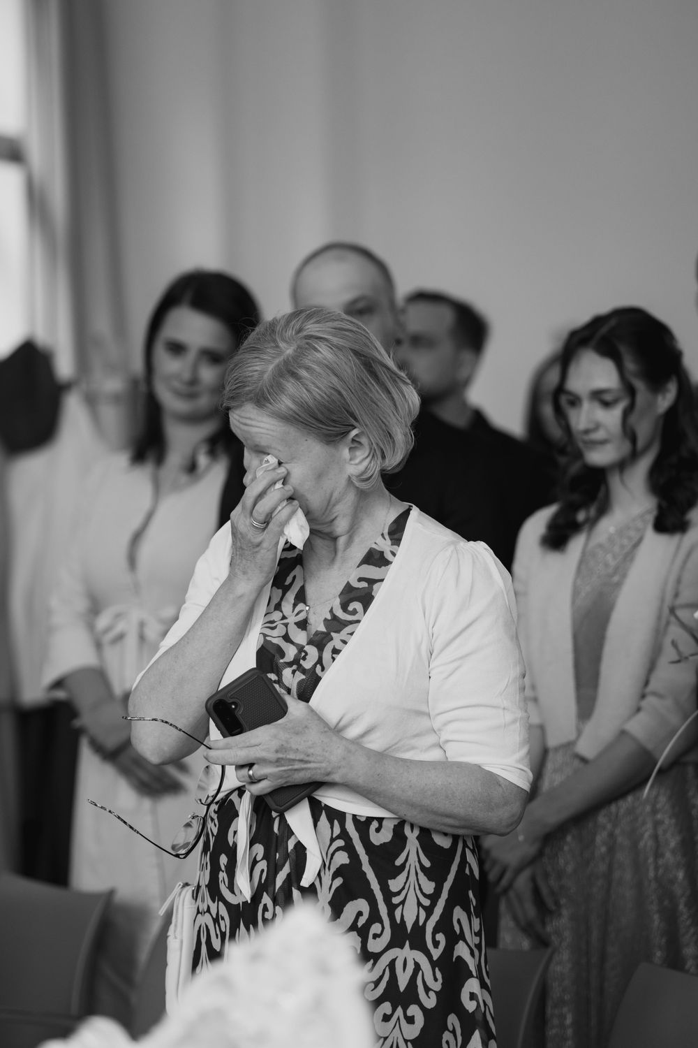 Black and white photo of an emotional moment at a gathering as someone in a white jacket and patterned dress wipes their eyes.