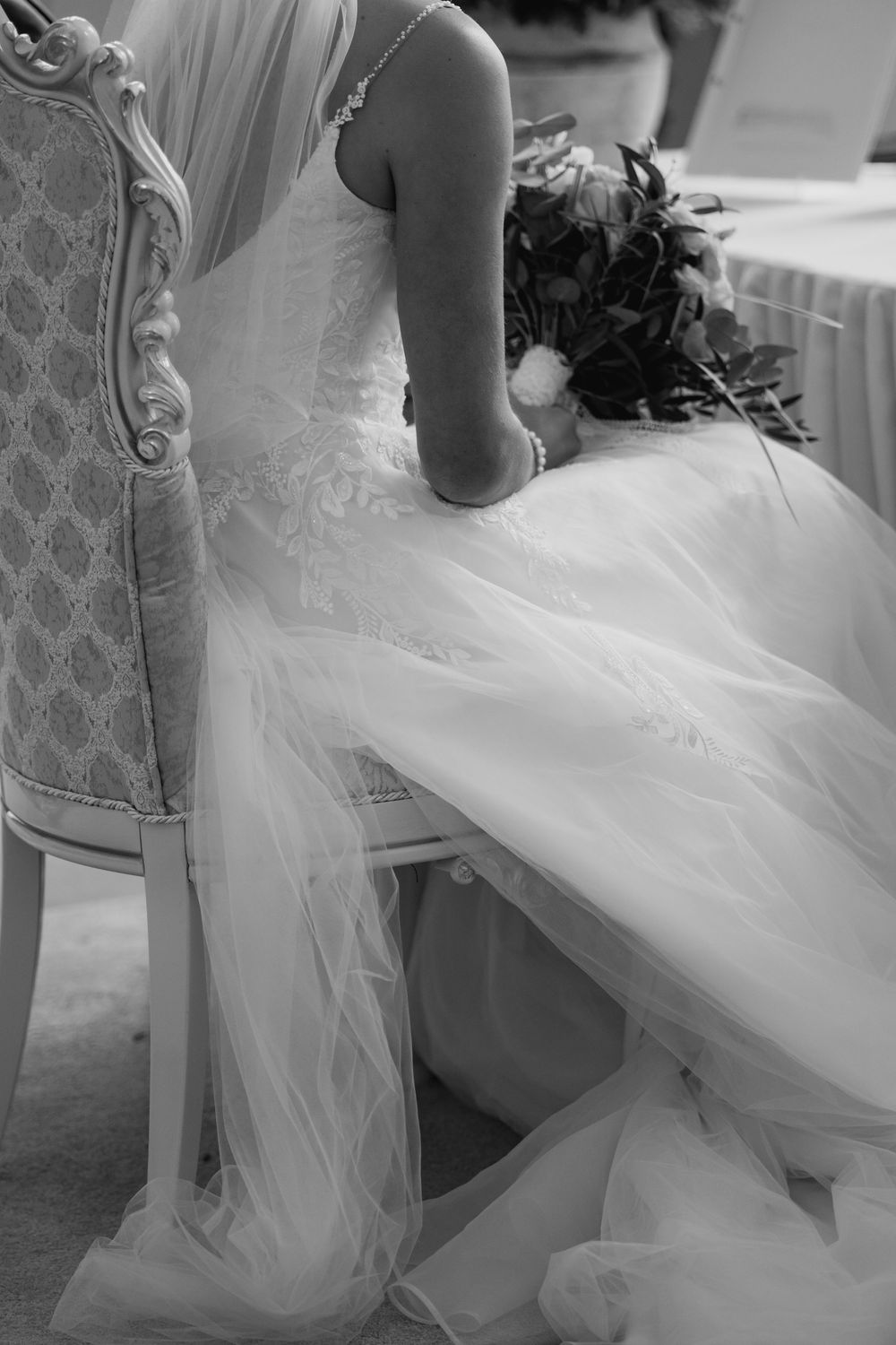 Black and white detail shot of a wedding dress with flowing tulle fabric draped over an elegant antique chair.