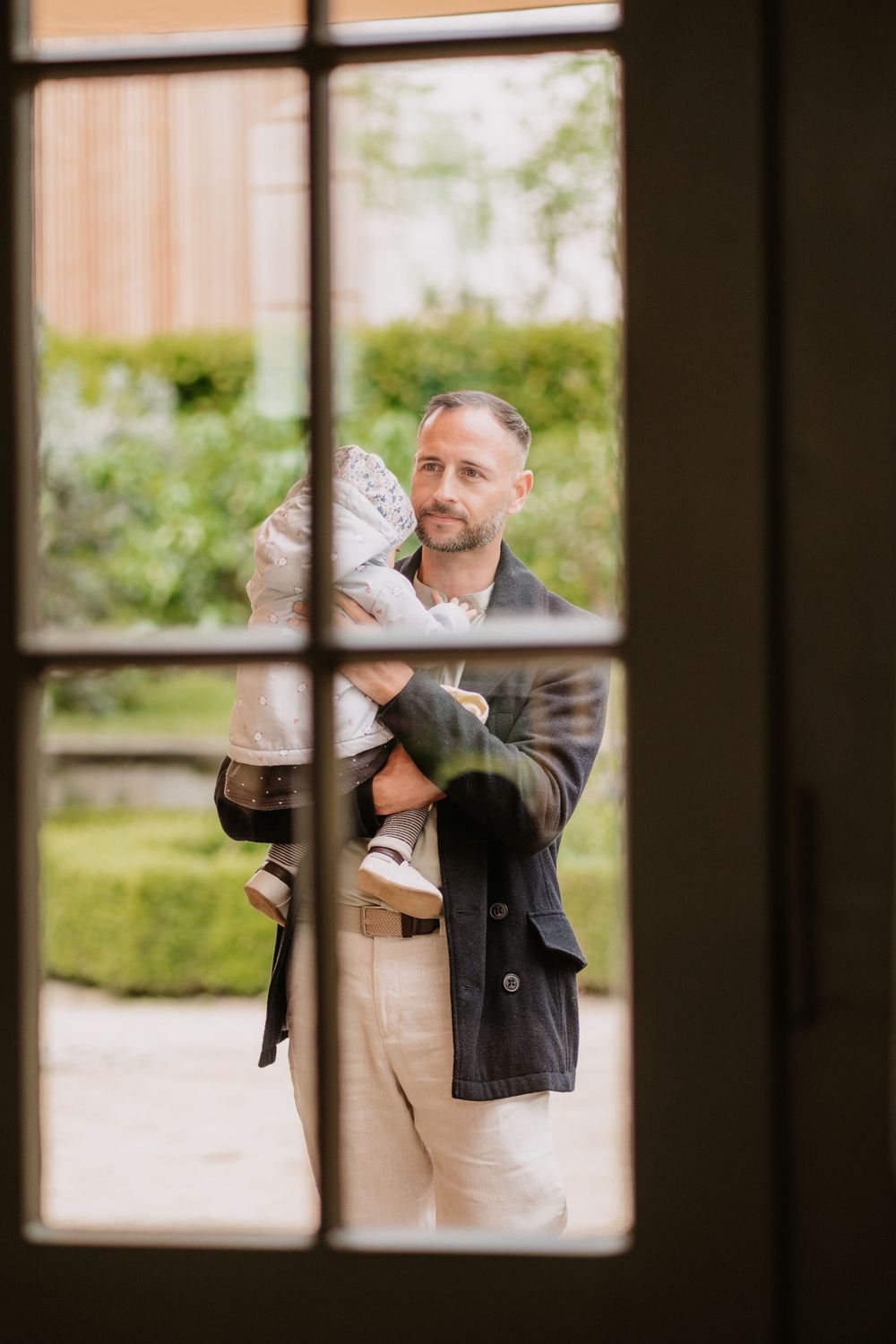 Through a windowpane, a tender moment is captured as someone in a suit holds a small child on a sunny day.