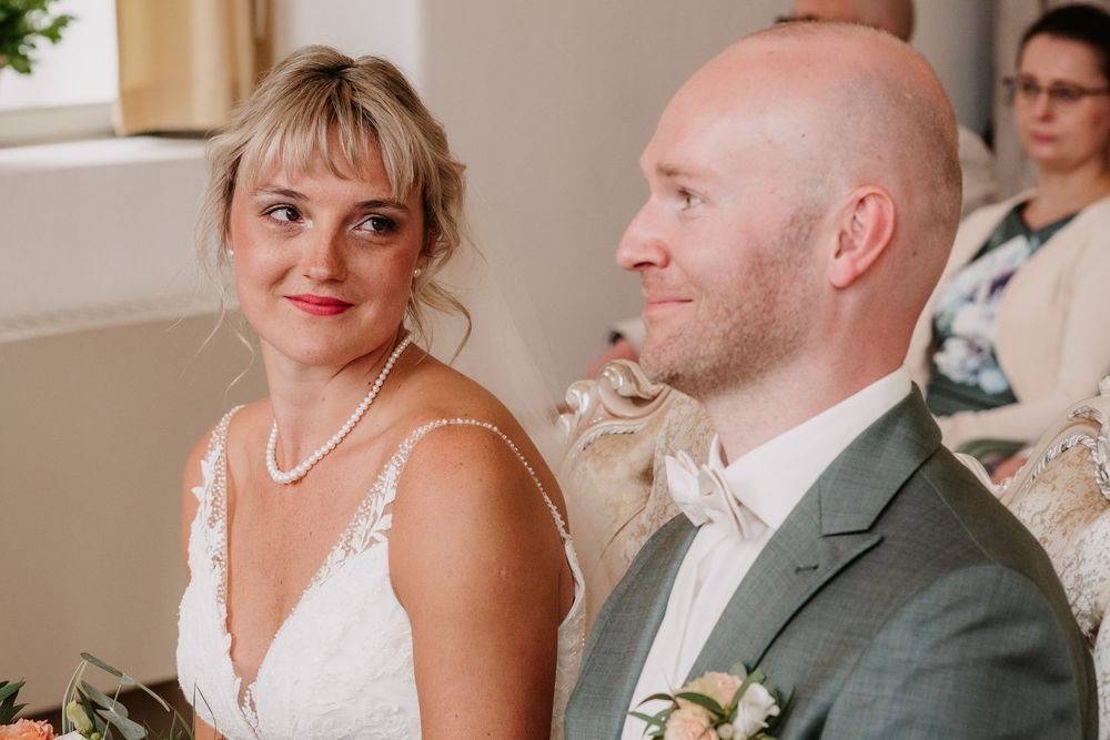 A couple at their wedding ceremony, with the bride wearing a white dress and pearl necklace, and groom in a gray suit.