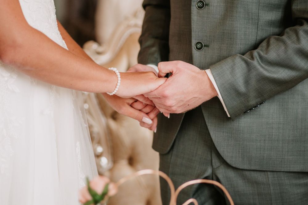Close-up view of a couple holding hands during a wedding ceremony, with the groom in grey suit and bride in white dress.
