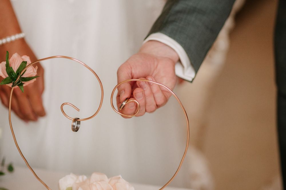 Close-up of hands holding delicate copper wire heart decoration with white dress fabric and greenery in background.