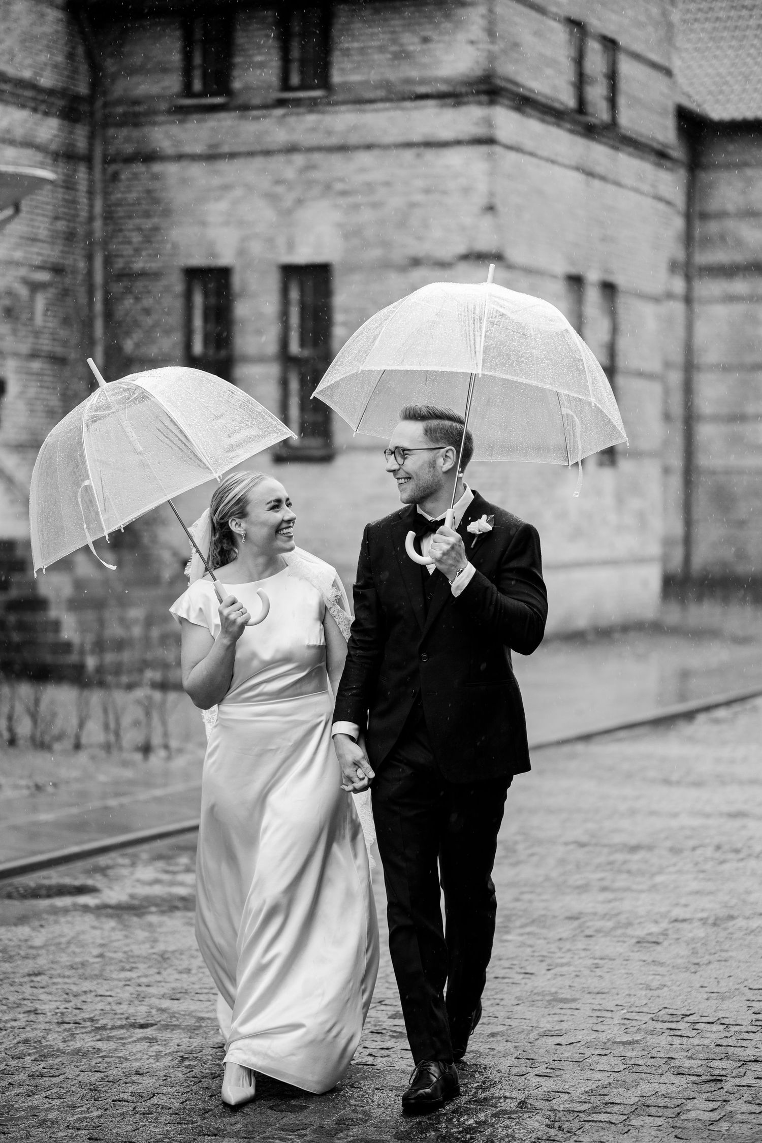 A couple in formal wedding attire stands together in a windswept field of tall grass by the ocean.