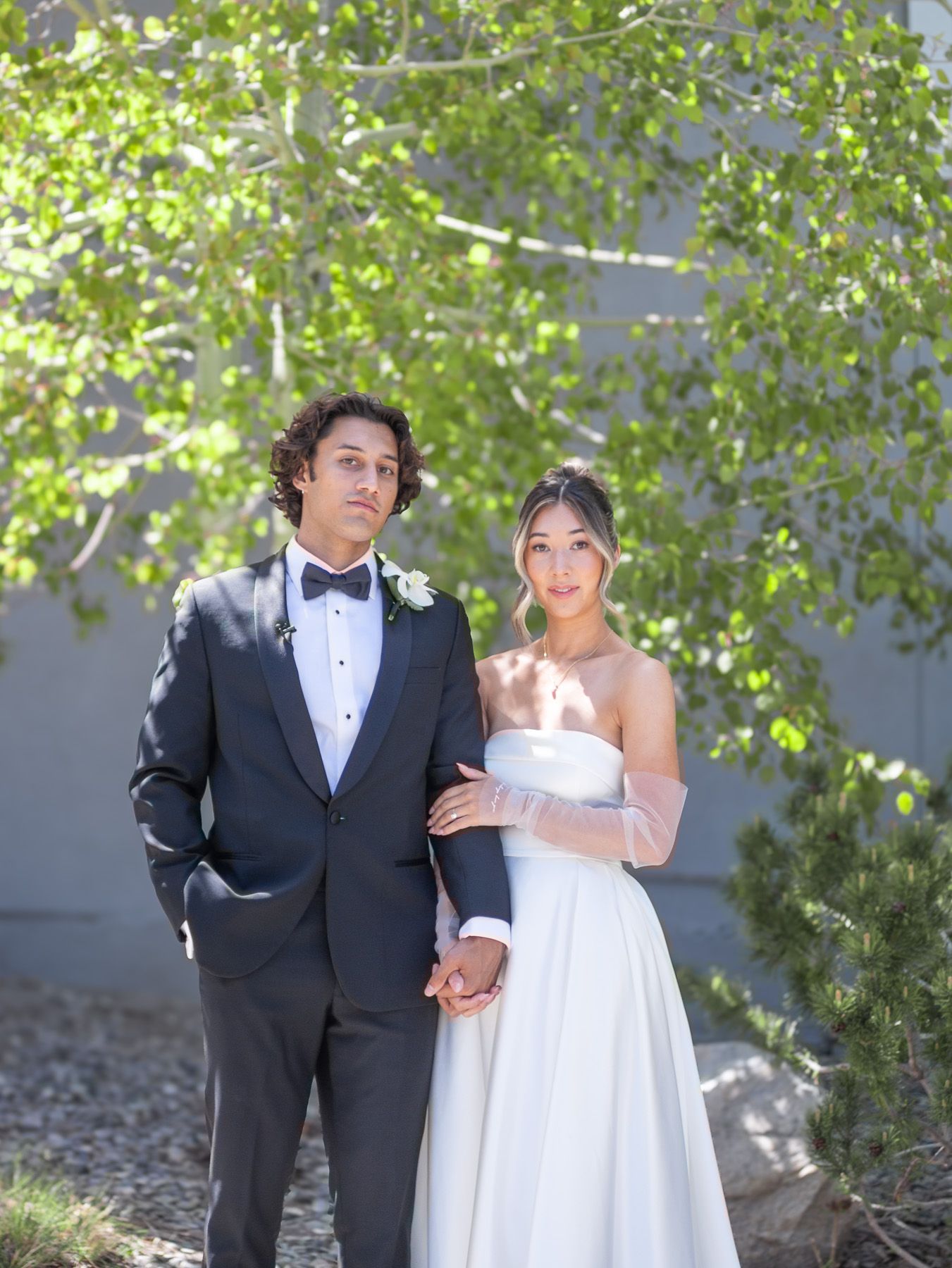 A couple in formal wedding attire poses outdoors under sunlit green trees on their special day.