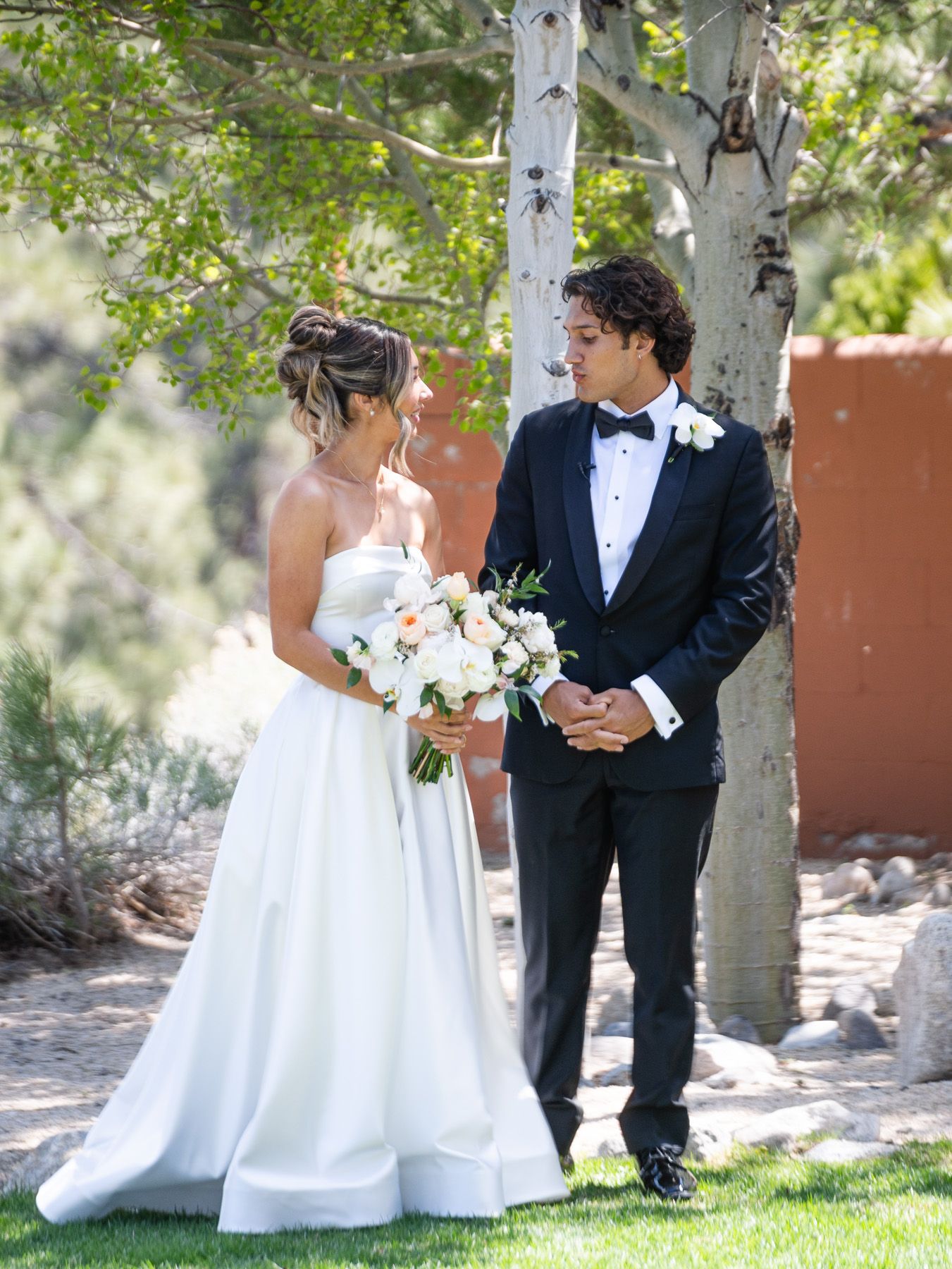 A romantic wedding couple stands together under birch trees, the bride holding a white and blush bouquet.