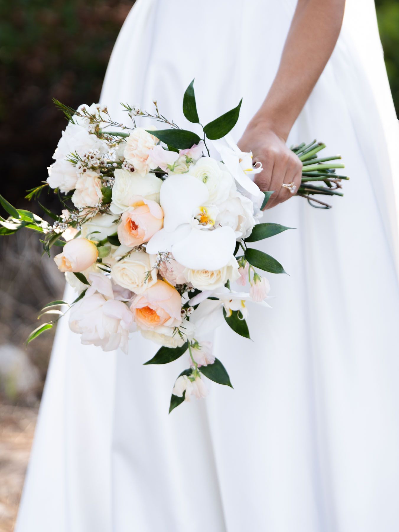 A romantic bouquet of peach and white peonies with greenery held against a white bridal gown in outdoor wedding setting.