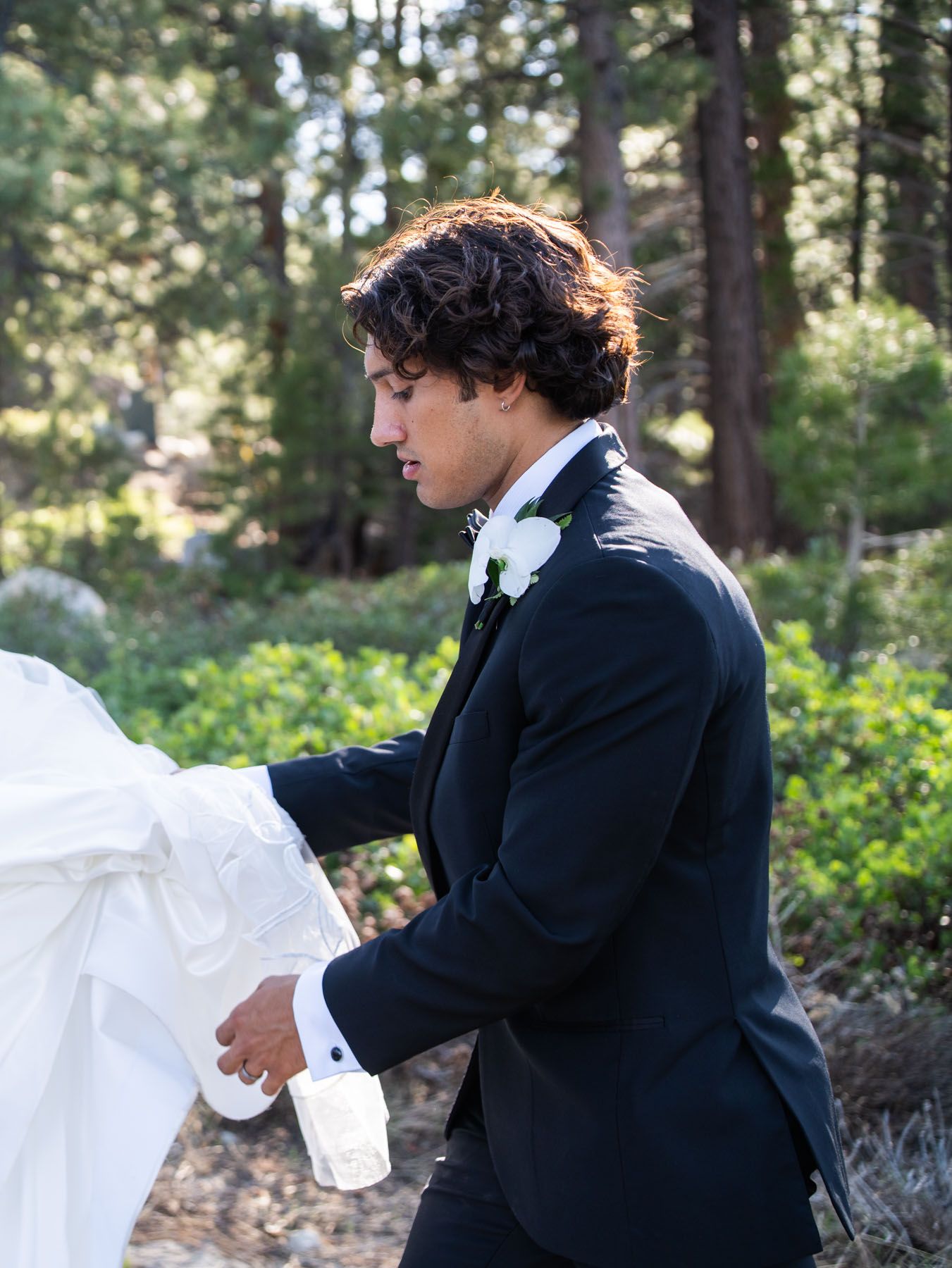 A groom in a black tuxedo with a boutonniere helps arrange a wedding dress in a sunlit forest setting.