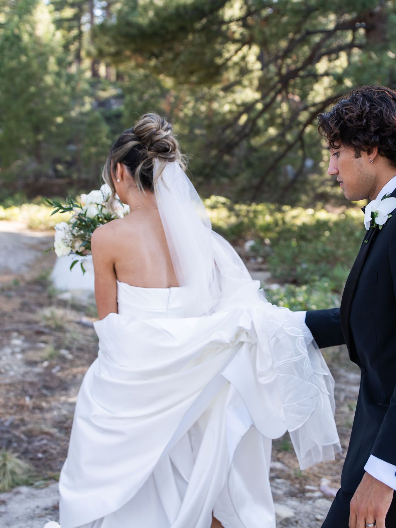 Bride in flowing white wedding gown holding bouquet walks through outdoor forest setting with groom in black suit.