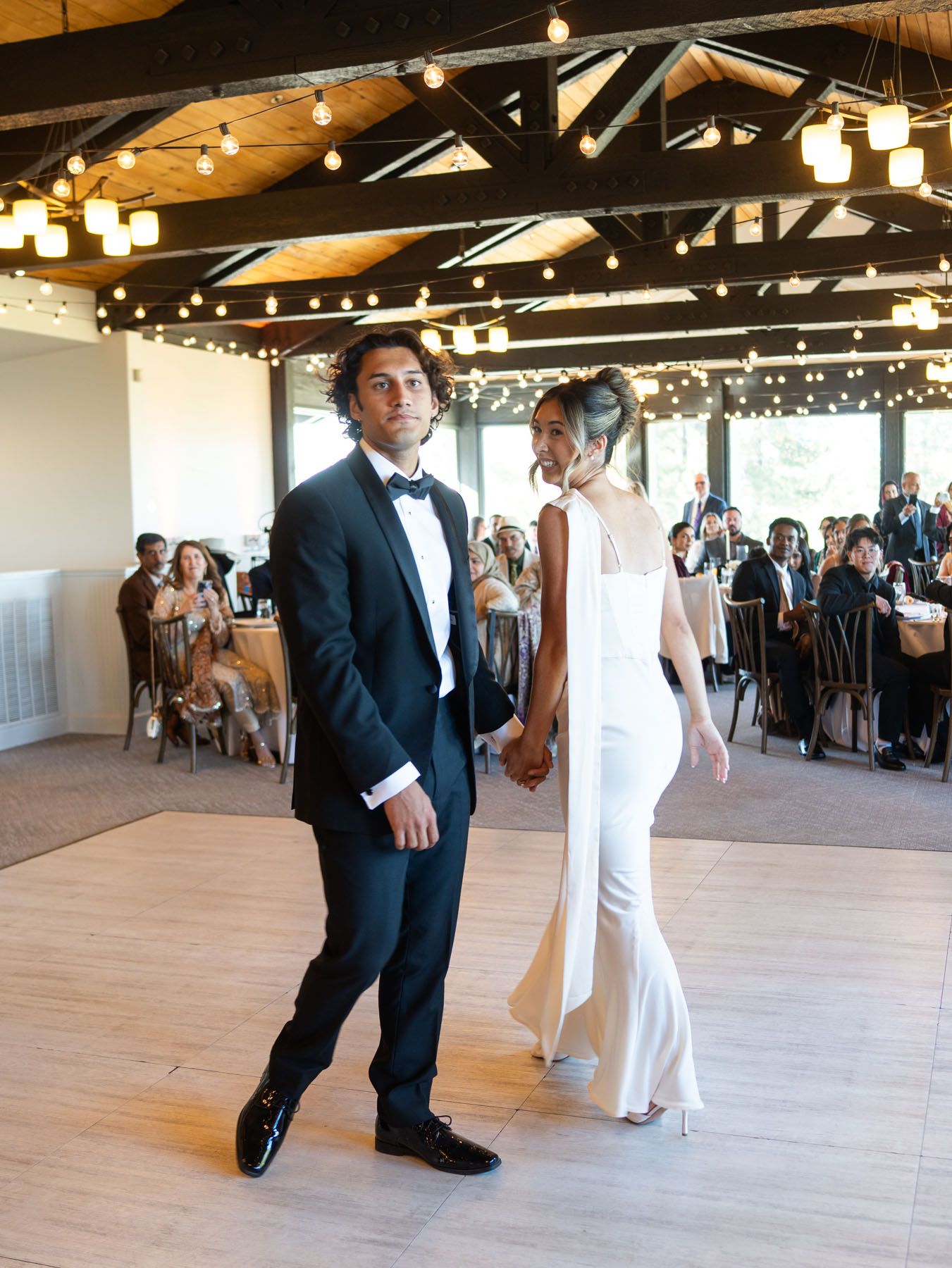 A couple shares their first dance in an elegant rustic venue decorated with string lights and exposed wooden beams.