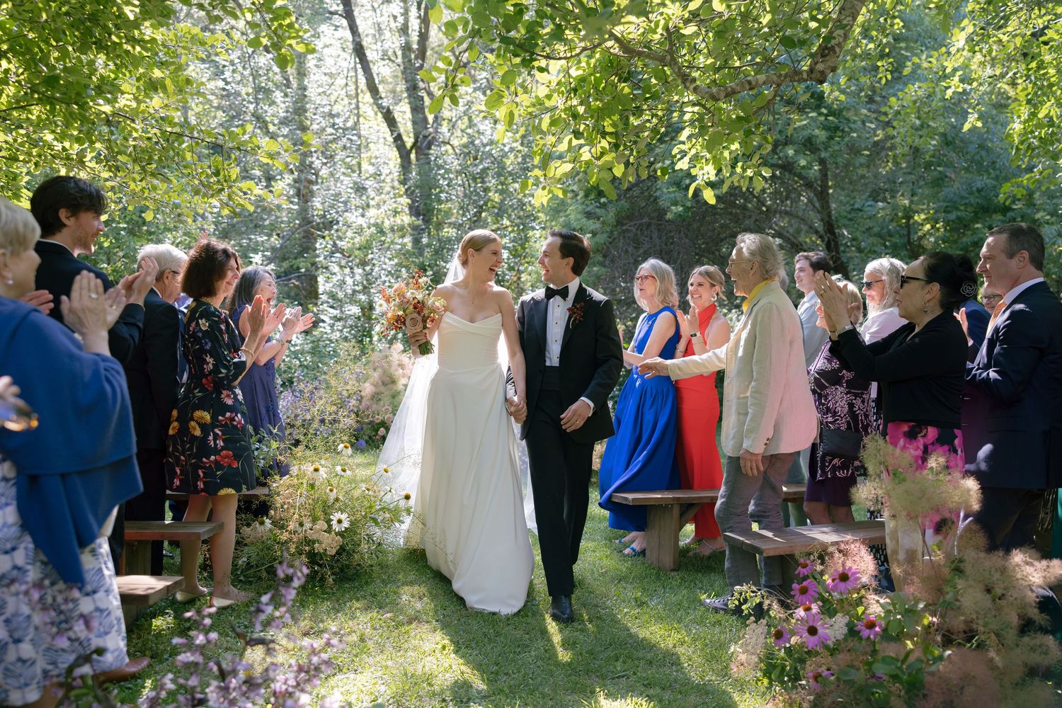 Newlyweds celebrate joyfully with guests during an outdoor forest wedding reception decorated with string lights.