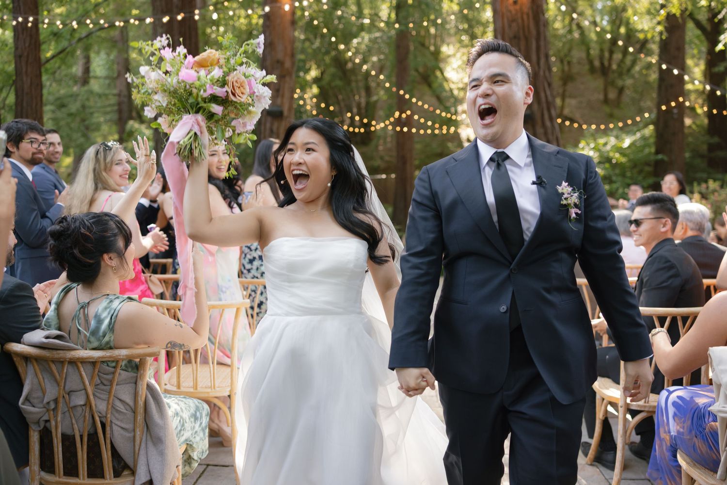 Newlyweds celebrate joyfully with guests during an outdoor forest wedding reception decorated with string lights.
