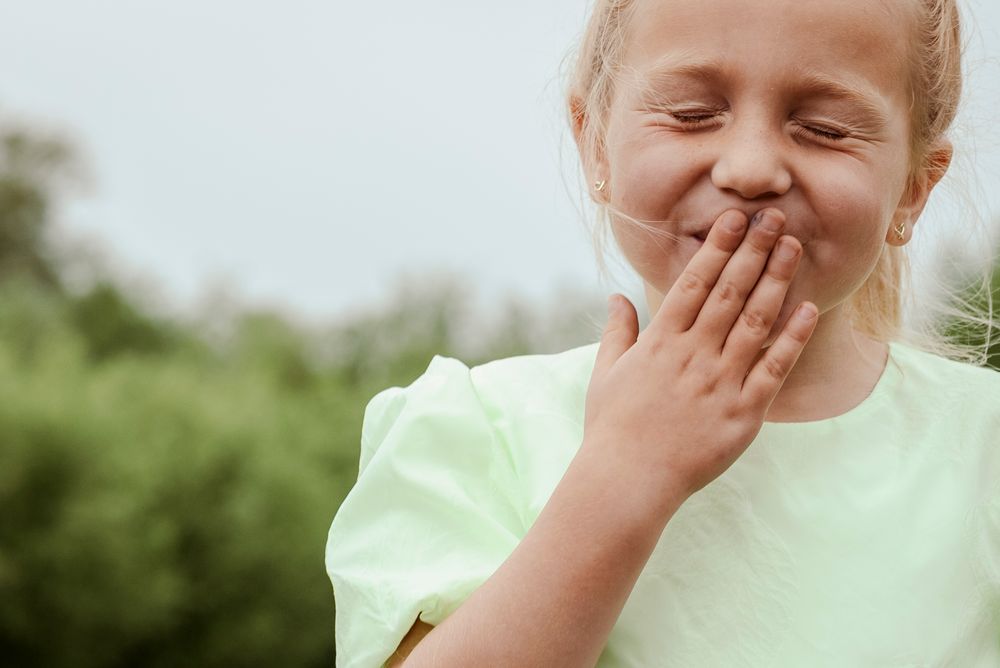 A young blonde girl covering her mouth and smiling with eyes closed outdoors in nature.
