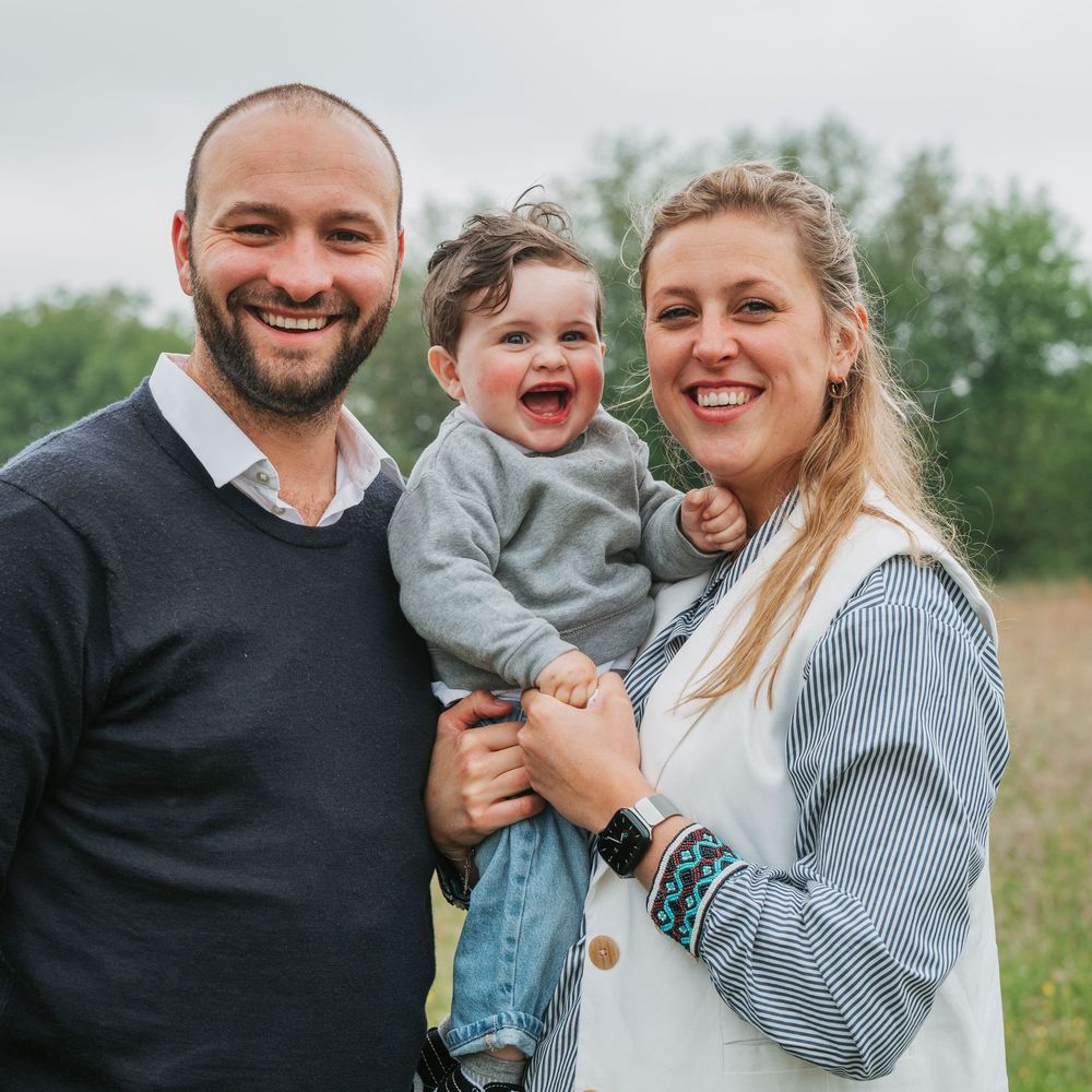 Happy young family with smiling baby boy posing outdoors in a green field.