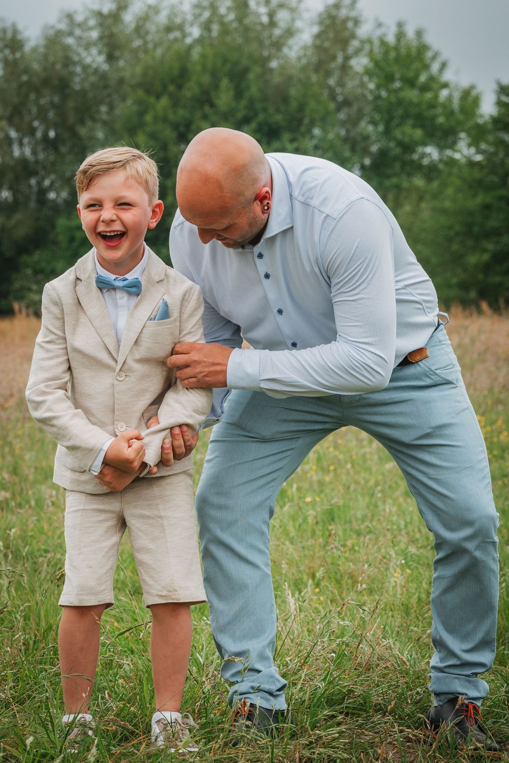 A bald father tickling his laughing son dressed in a linen suit with a blue bow tie outdoors.