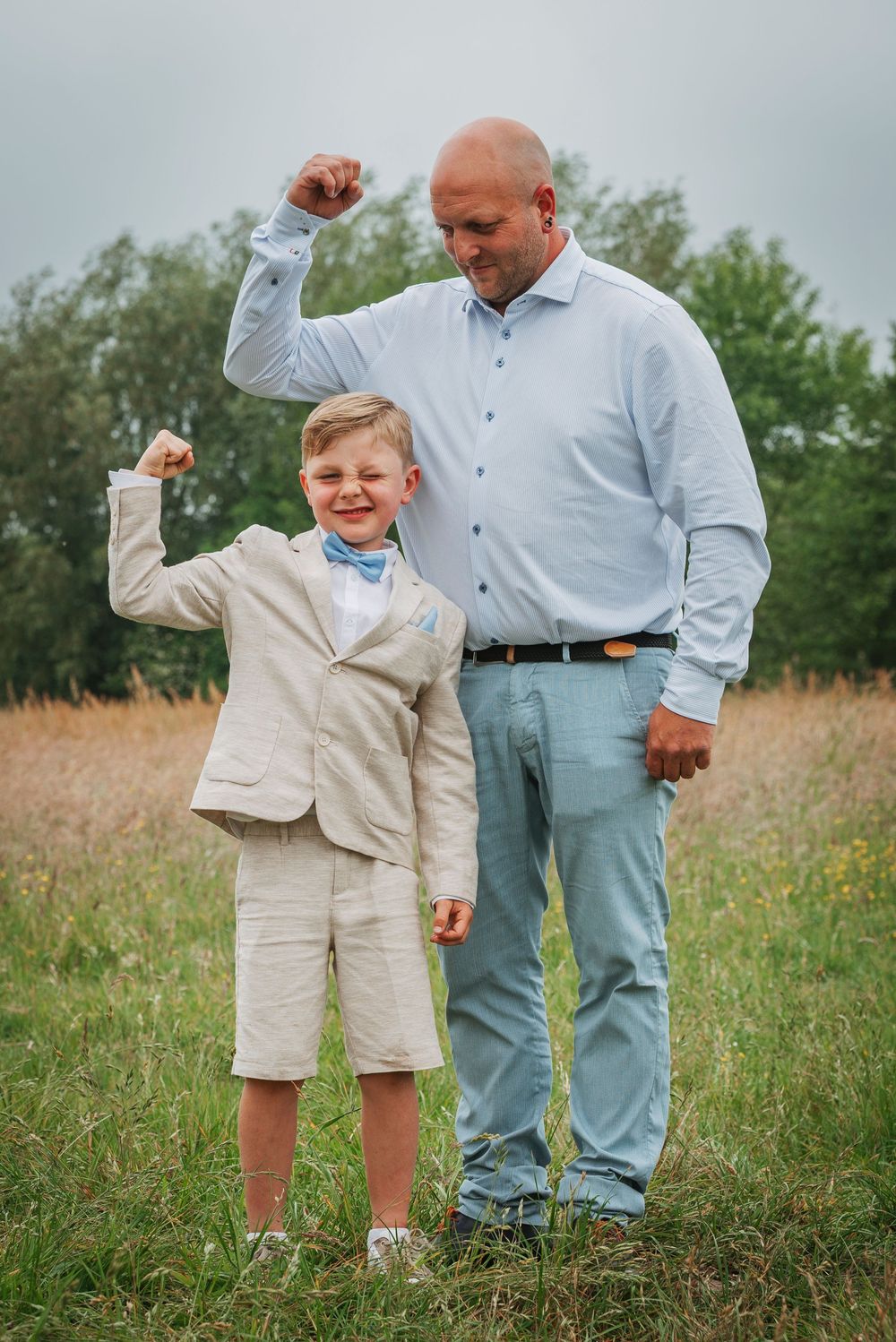 A bald father and young son in a suit flexing their muscles together in a green field.