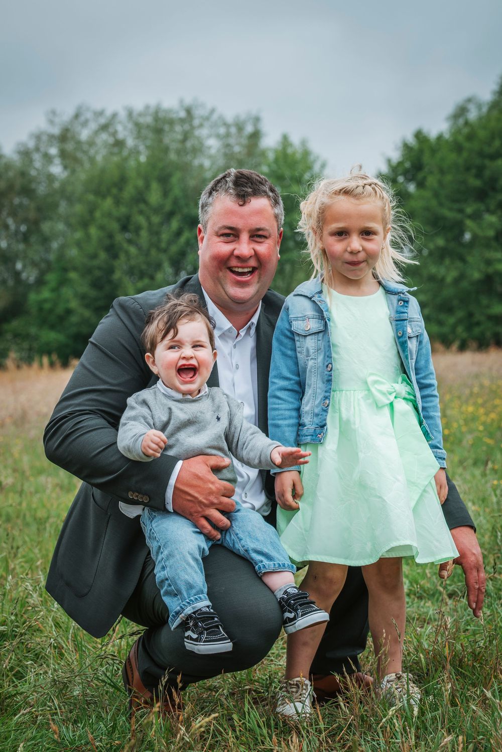 Smiling man crouching with laughing baby boy and young blonde girl in field.