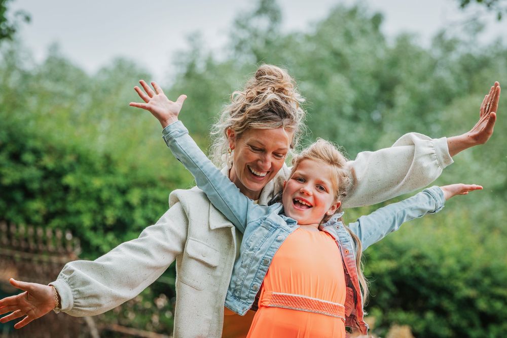 Joyful mother and daughter spreading arms wide outdoors, laughing together in a garden.