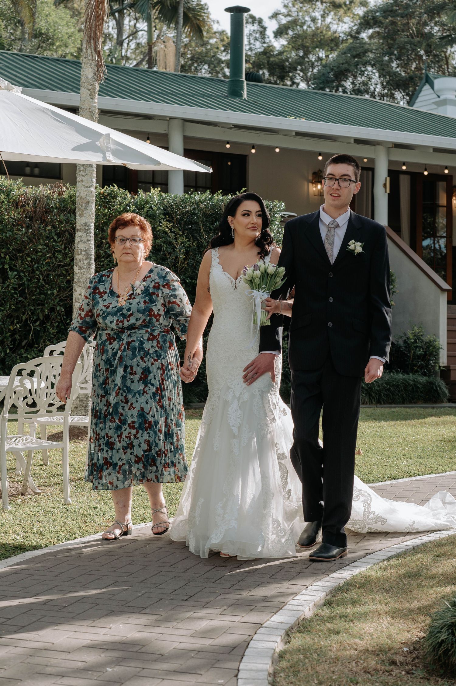 Wedding procession walking down a paved path outside a venue with a green roof and white columns on a sunny day.