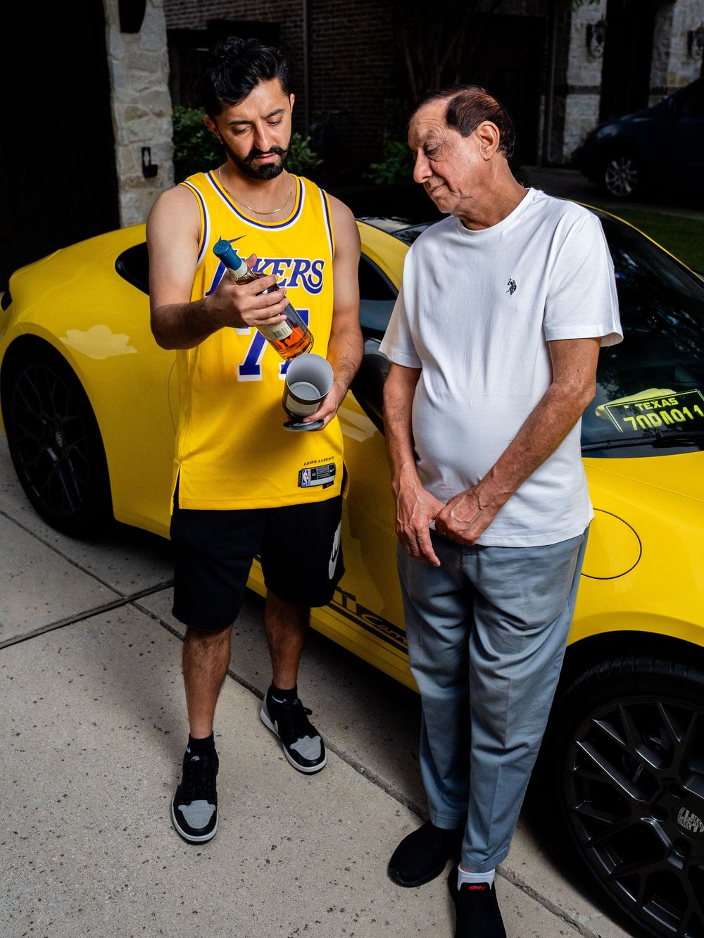Two men stand next to a bright yellow sports car, one wearing a Lakers jersey and one in a white t-shirt.