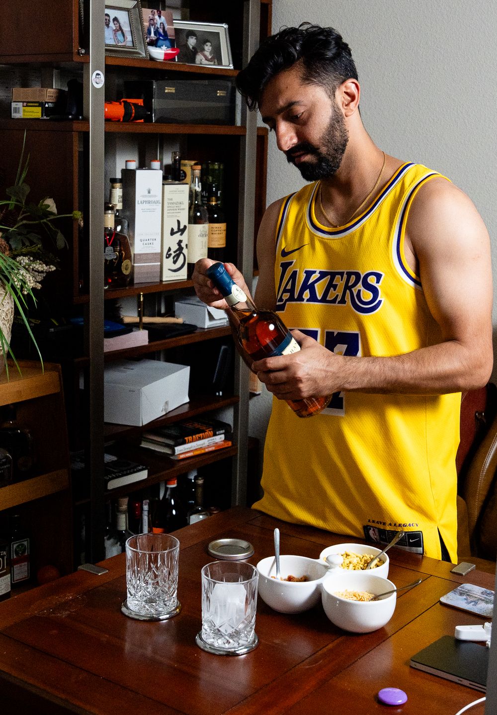 Person in Lakers jersey pouring cocktail ingredients at home bar setup with vintage glassware on wooden table.