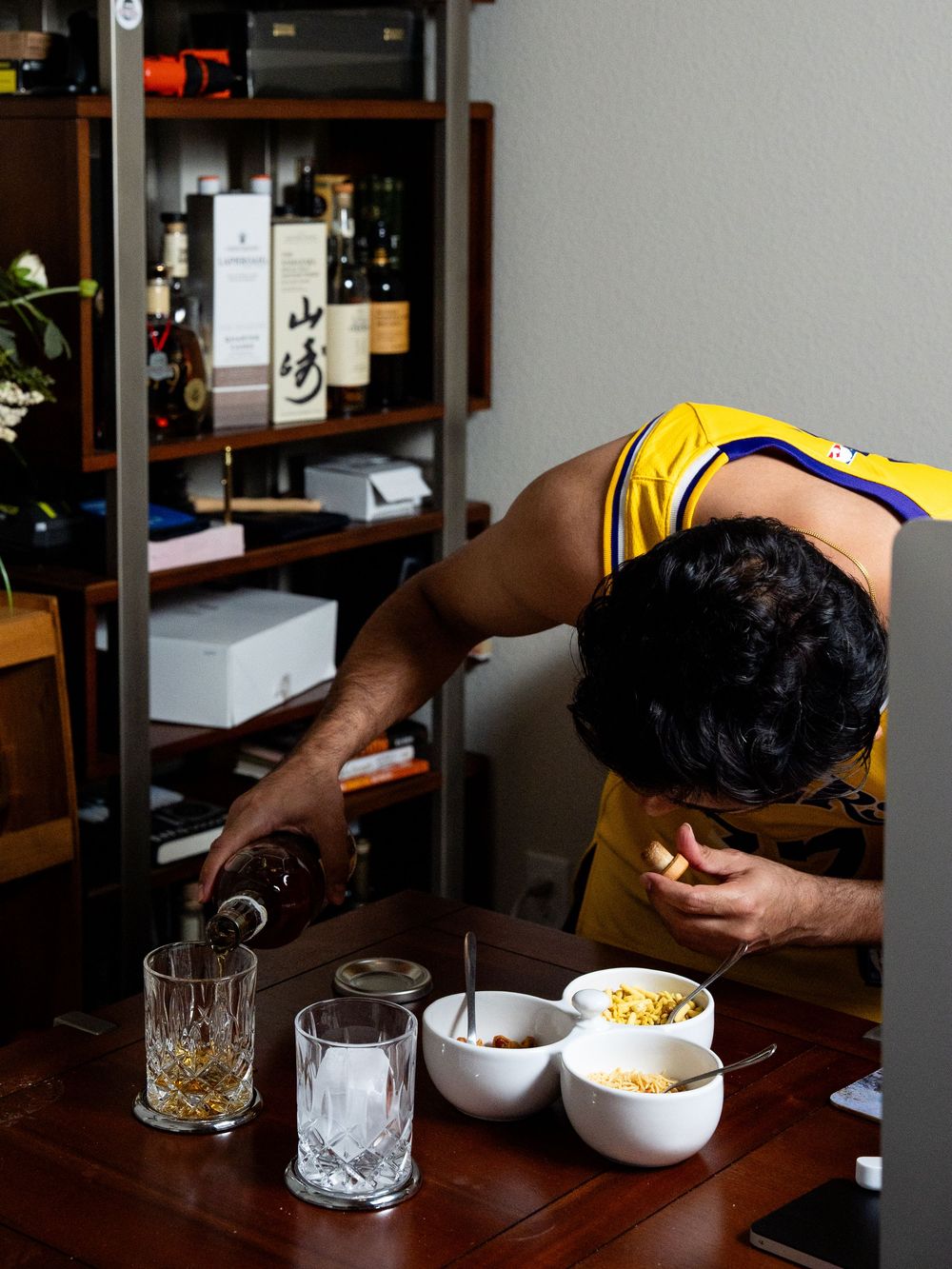 Person in yellow shirt pouring a drink beside bowls of snacks and crystal glasses on a wooden table.