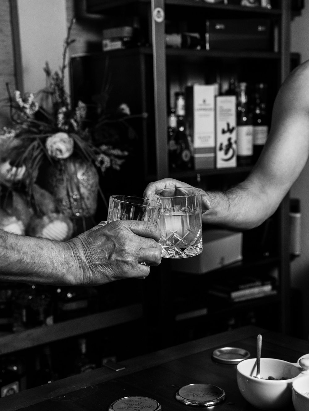 Black and white photo of two hands exchanging a glass of whiskey with bottles and bar supplies in the background.