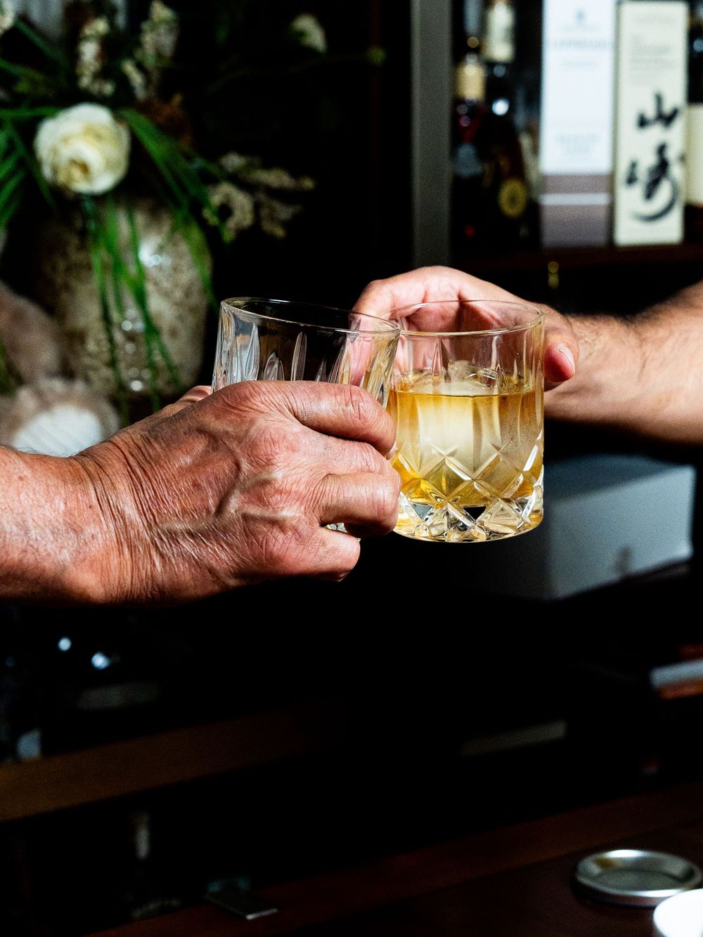 A weathered hand holding a glass of whiskey with ice cubes at a dark wooden bar counter.