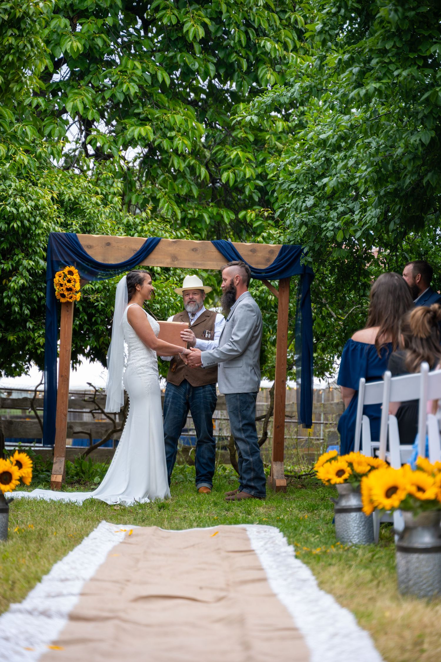 Couple exchanges vows under rustic wooden arch with navy draping and sunflowers at outdoor garden wedding ceremony.