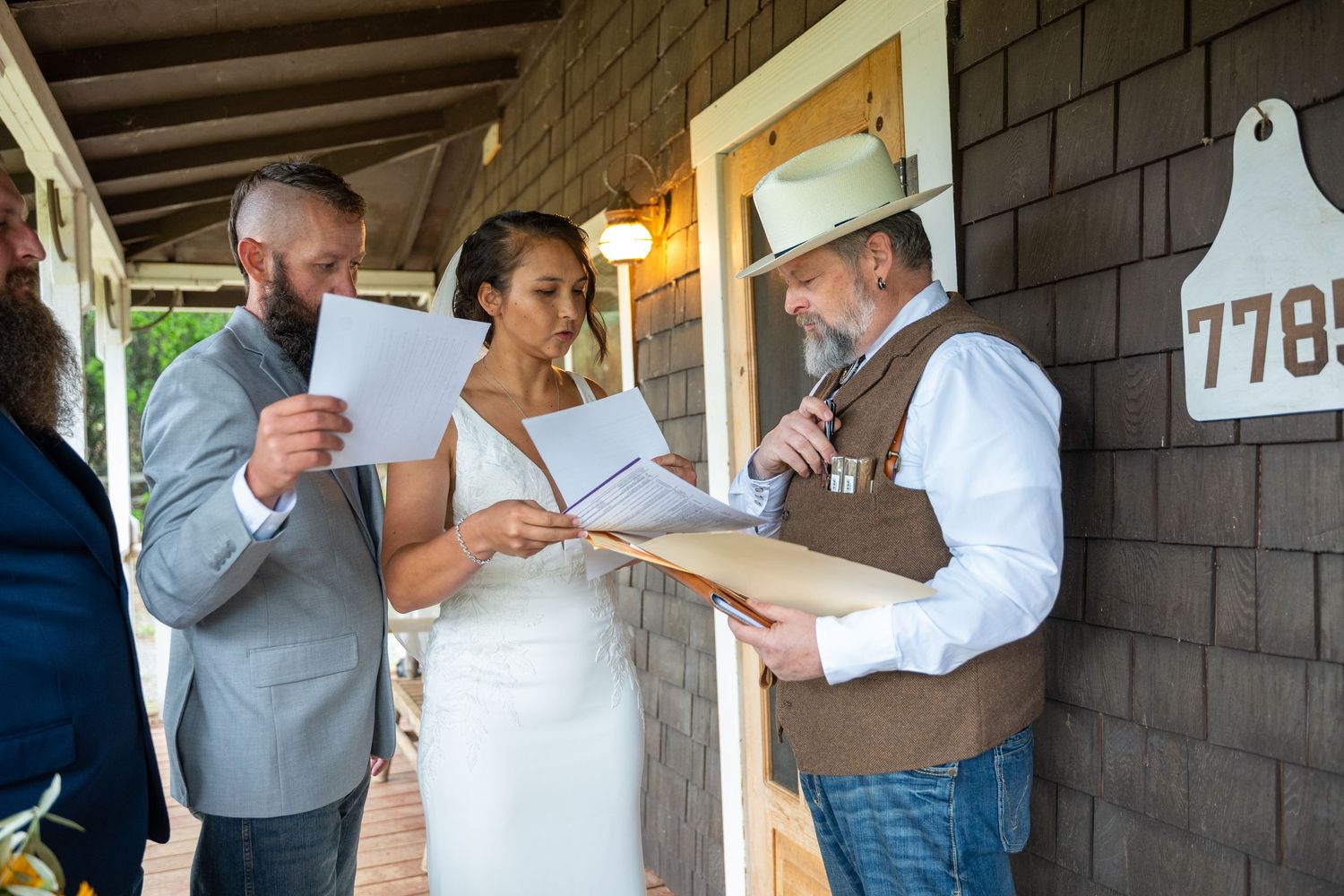 Bride and groom signing wedding documents with officiant in cowboy hat on rustic porch.