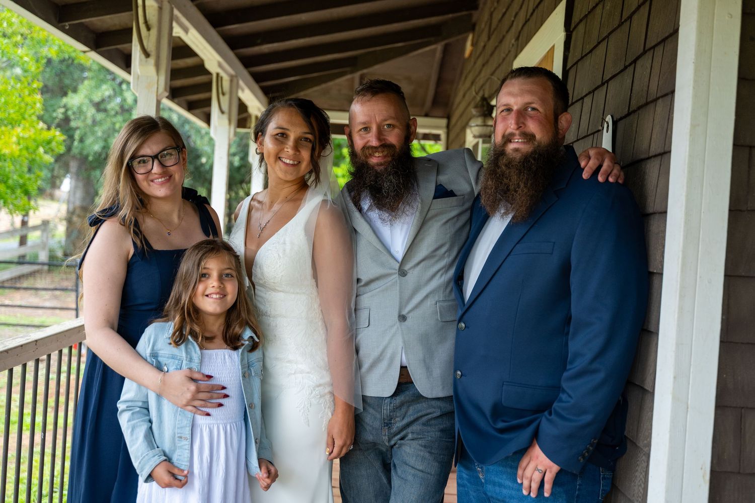 Family group poses in wedding attire on a porch, featuring a bride in white gown with bearded groom in gray suit.